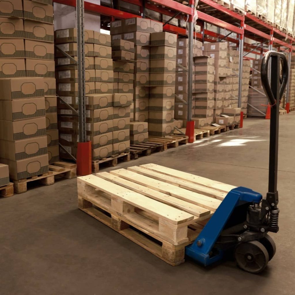Wooden pallet handled with a manual pallet jack inside a warehouse with stacked goods on pallet racking systems